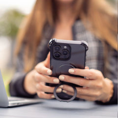 Person holding an iPhone 16 Plus with a Raptic iPhone 16 Shield Impact Case - Dual Impact and a ring grip beside a laptop outdoors, showcasing the case's drop protection features and MagSafe compatibility.