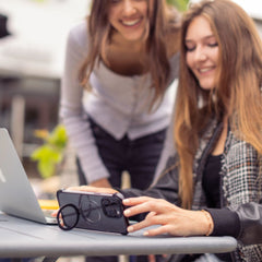 Two women are at a table outside. One, sitting and using a smartphone equipped with the Raptic iPhone 16 Shield Impact Case - Dual Impact for drop protection, is near an open laptop. The other is standing and smiling, bending slightly towards the table.