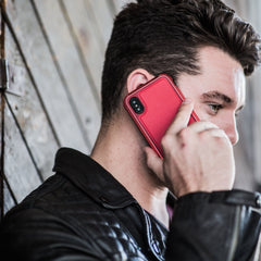 A person with short dark hair holds a red smartphone in an impact-resistant Raptic LUX case for the iPhone XS Max to their ear, wearing a black leather jacket and standing against a wooden wall.