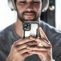 A man with finger tattoos holds an iPhone 11 Pro, protected by a Raptic SHIELD case, in both hands. He wears headphones and a gray shirt in a dimly lit room.