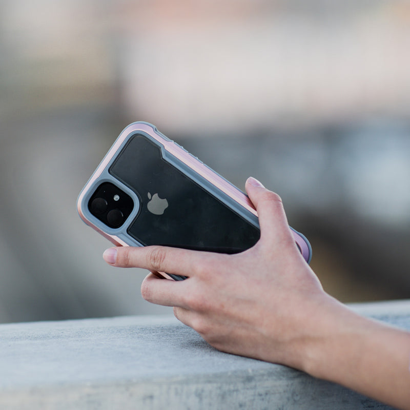 A hand holding an iPhone 11 Pro with a dual-camera setup and an Apple logo, encased in a Raptic SHIELD clear protective case made from aluminum alloy, outdoors.