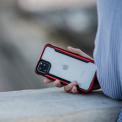 A person holds an iPhone 11 Pro encased in a Raptic SHIELD case, featuring a red and black aluminum alloy design, and rests it on an outdoor concrete surface.