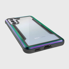 An X-Doria Samsung Galaxy S21 Raptic Shield Iridescent case highlights dual rear cameras, offers military-grade protection, a green and purple iridescent border with a black bottom edge, and remains wireless charging compatible.