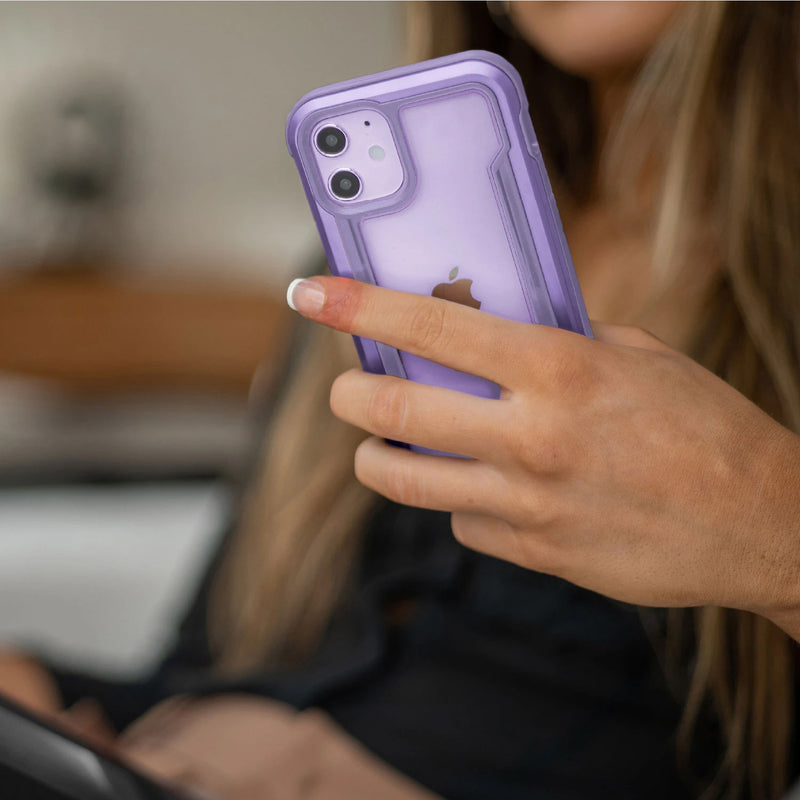 Indoors, a person holds an iPhone 12 with a Raptic Shield case in purple. The sleek machined aluminium bumper offers military-grade drop protection and wireless charging compatibility, blending style with peace of mind.