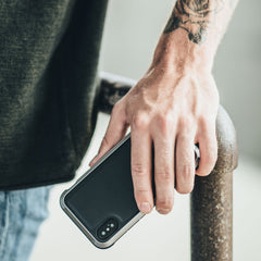 A tattooed individual leans casually on a metal railing, holding a smartphone adorned in the Raptic LUX iPhone XR Case. The impact-resistant polycarbonate shell, enhanced with bubble-patterned rubber, offers both style and protection to their daily tech companion.