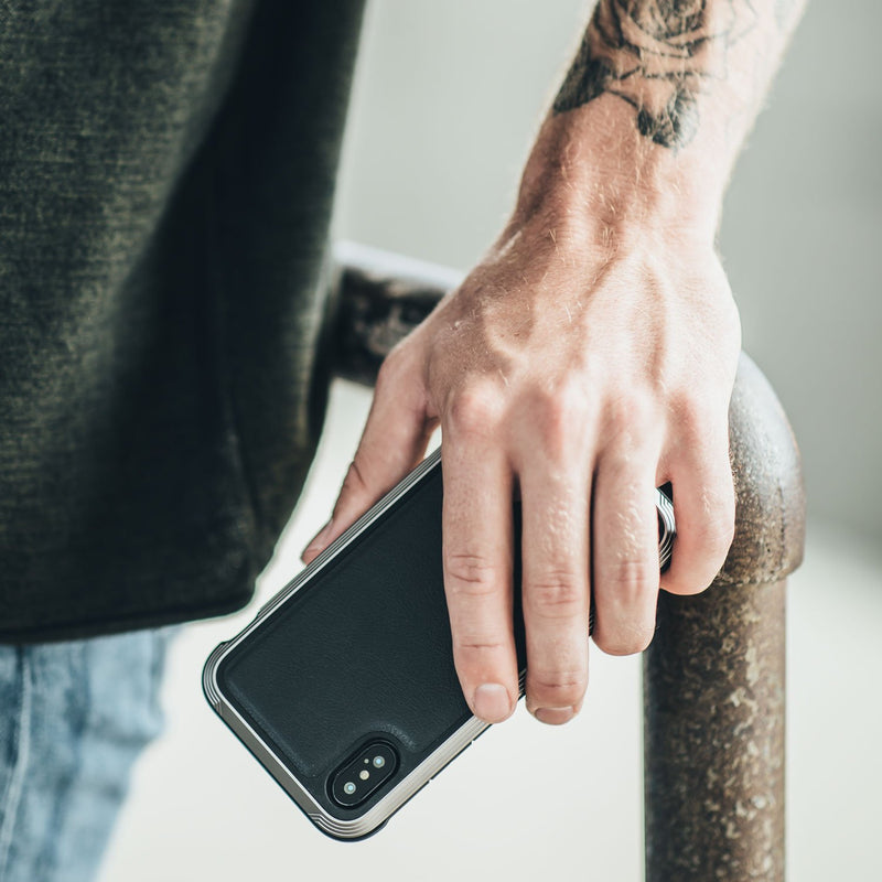 A tattooed individual leans casually on a metal railing, holding a smartphone adorned in the Raptic LUX iPhone XR Case. The impact-resistant polycarbonate shell, enhanced with bubble-patterned rubber, offers both style and protection to their daily tech companion.