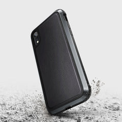 A Raptic LUX black iPhone XR case with a bubble-patterned rubber surface stands on edge, surrounded by dust and debris on the cracked gray ground.