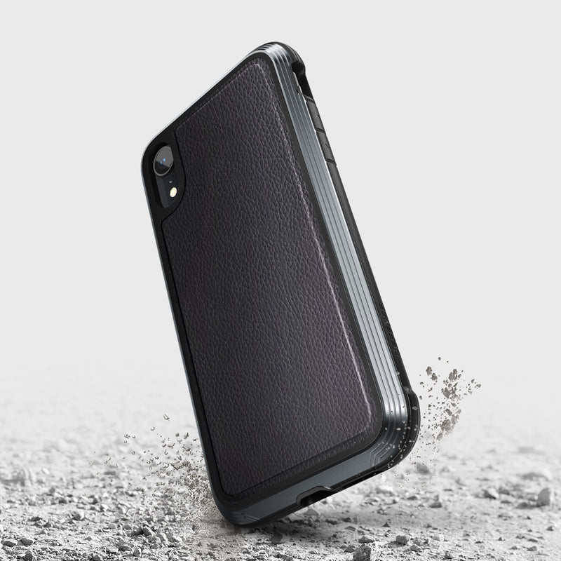 A Raptic LUX black iPhone XR case with a bubble-patterned rubber surface stands on edge, surrounded by dust and debris on the cracked gray ground.