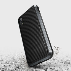 The Raptic iPhone XR Case - LUX, featuring black carbon fiber texture with impact-resistant polycarbonate and rugged edges, is showcased on a cracked concrete surface to highlight its durability.