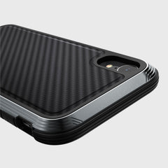 A close-up of the Raptic LUX iPhone XR case showcases its black carbon fiber textured surface, highlighting the rear camera area. The durable impact-resistant polycarbonate shell stands out against a plain backdrop.