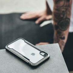 A tattooed person in the blurred background watches as an iPhone XR, secured in a Raptic SHIELD military-grade case, withstands a drop on concrete.