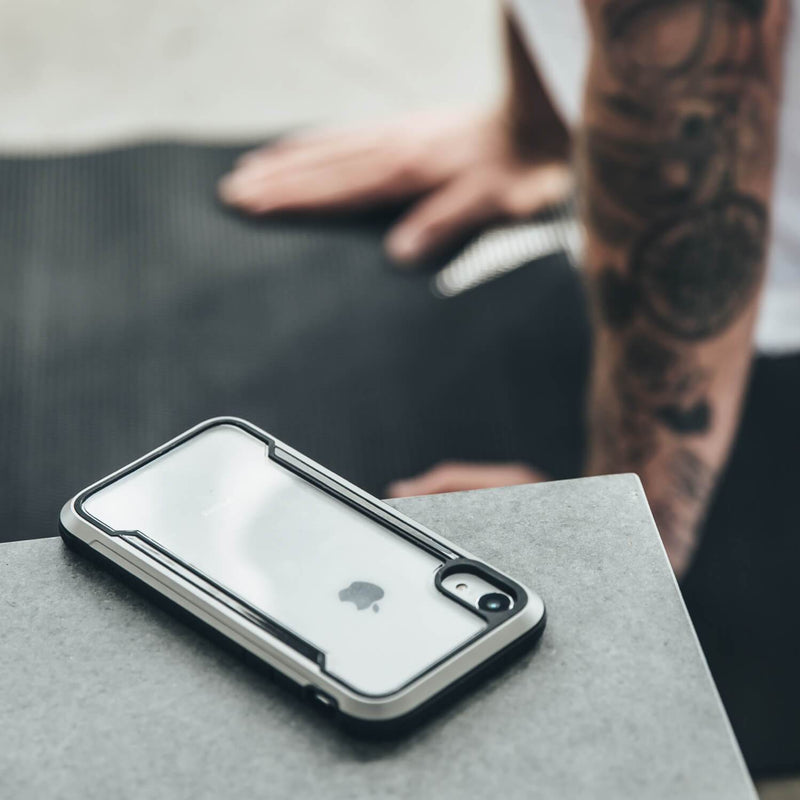A tattooed person in the blurred background watches as an iPhone XR, secured in a Raptic SHIELD military-grade case, withstands a drop on concrete.