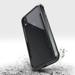 An iPhone XR in a rugged black Raptic SHIELD case with military-grade drop protection stands upright on a surface with small debris around it.
