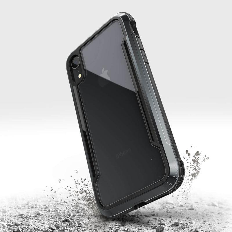 An iPhone XR in a rugged black Raptic SHIELD case with military-grade drop protection stands upright on a surface with small debris around it.