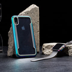 An iPhone XR in a colorful Raptic SHIELD case with military-grade drop protection and shock absorption is displayed with a smartwatch featuring a gray band against concrete blocks on a dark surface.