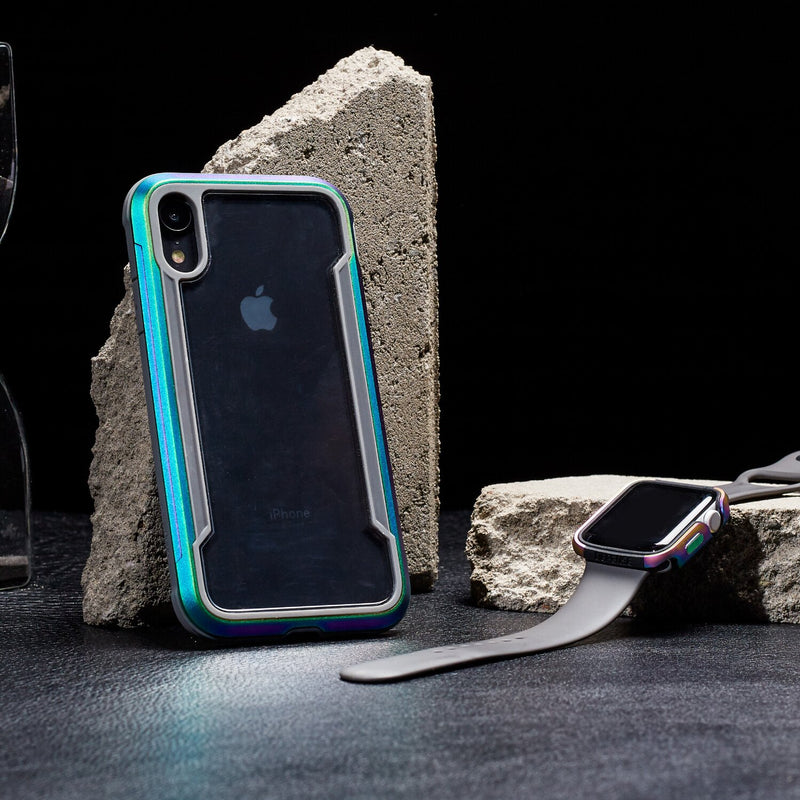 An iPhone XR in a colorful Raptic SHIELD case with military-grade drop protection and shock absorption is displayed with a smartwatch featuring a gray band against concrete blocks on a dark surface.