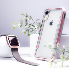 An iPhone XR in a pink and gray Raptic SHIELD case, designed for military-grade drop protection, is positioned upright next to a smartwatch with a dark band. Both rest stylishly among small potted plants on a pristine white surface.