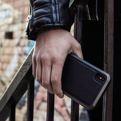 A person holds a Raptic iPhone XS Max Case - LUX in black while on stairs, sporting a black leather jacket. Its sleek design complements the look, and the impact-resistant polycarbonate offers durability.