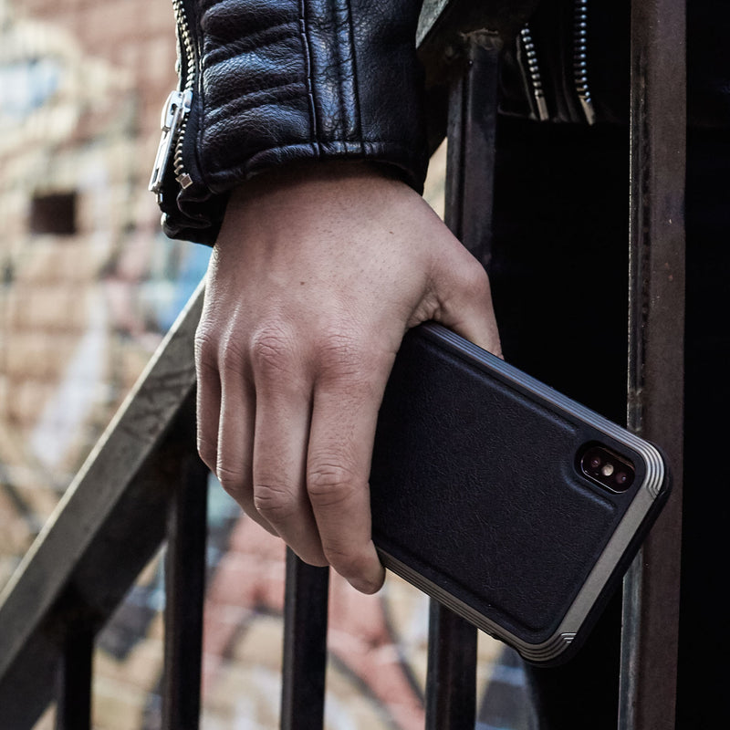 A person holds a Raptic iPhone XS Max Case - LUX in black while on stairs, sporting a black leather jacket. Its sleek design complements the look, and the impact-resistant polycarbonate offers durability.
