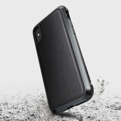 The Raptic iPhone XS Max Case - LUX, made from an impact-resistant polycarbonate, is shown from the back in a rugged black design. It sits on a textured surface with small debris, highlighting its durability.