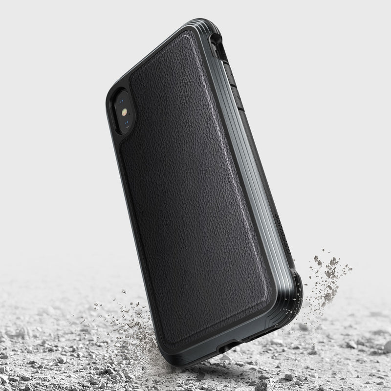 The Raptic iPhone XS Max Case - LUX, made from an impact-resistant polycarbonate, is shown from the back in a rugged black design. It sits on a textured surface with small debris, highlighting its durability.