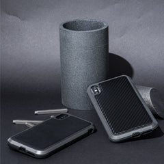 Two smartphones in rugged, impact-resistant polycarbonate cases are placed next to a dark cylindrical object and metal rods on a textured surface, showcasing the sleek design and durability of the iPhone XS Max Case - LUX by Raptic against machined aluminum alloy.