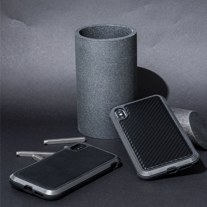 Two smartphones in rugged, impact-resistant polycarbonate cases are placed next to a dark cylindrical object and metal rods on a textured surface, showcasing the sleek design and durability of the iPhone XS Max Case - LUX by Raptic against machined aluminum alloy.