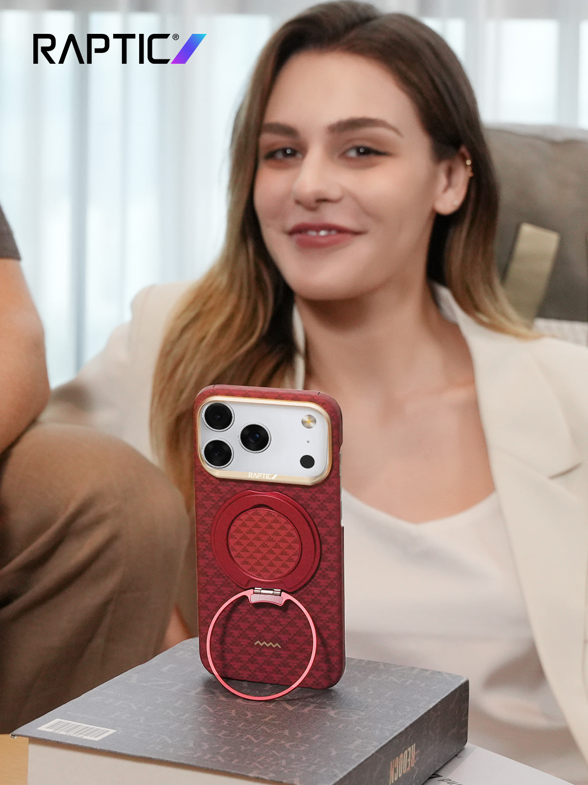 A woman sits behind a smartphone in a red case with a ring stand, positioned on a book. The RAPTIC logo is visible in the top left corner.