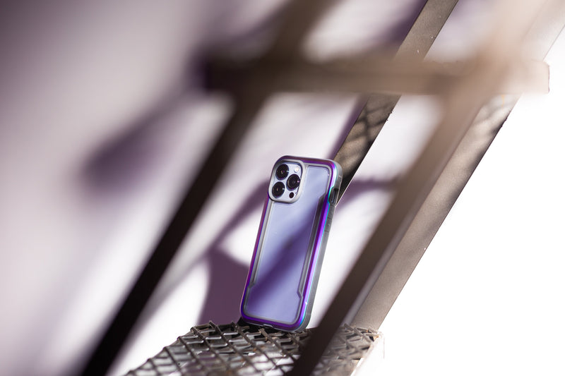 A smartphone with a purple case is propped against a metal structure. The phone's camera lenses are prominently visible and protected with a Raptic case