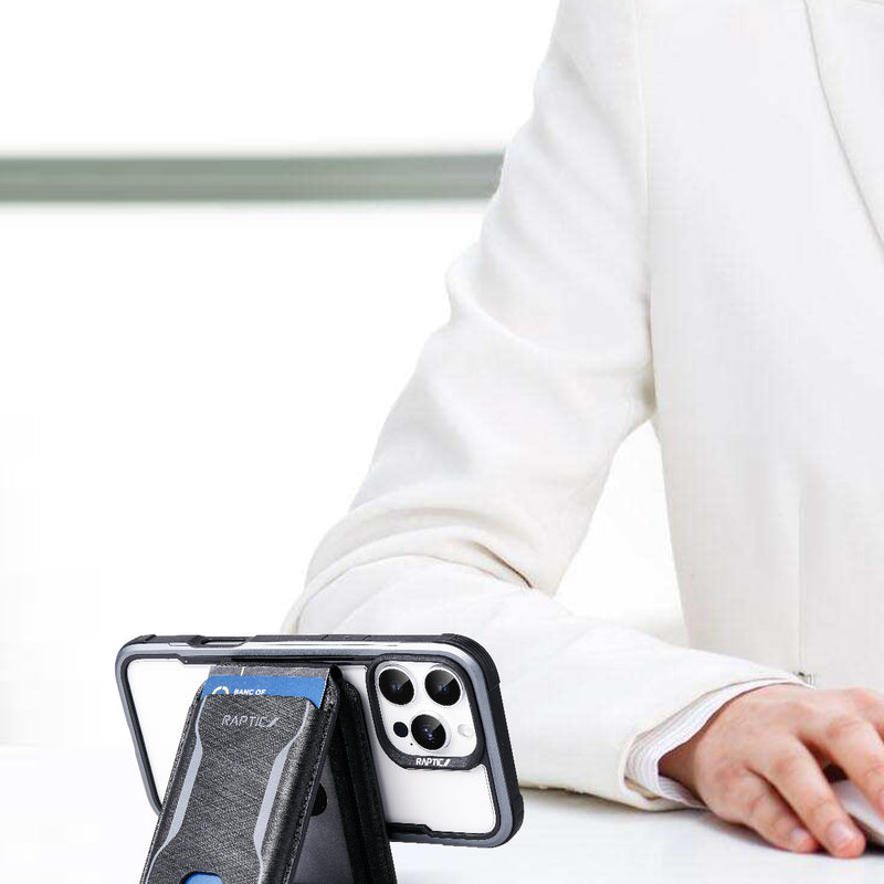A person in a white suit sits at a desk with a smartphone in a black Raptic Air Wallet case and a MagSafe Stand positioned in the foreground.