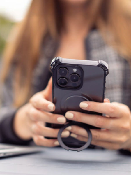 Person holding an iPhone 16 Plus with a Raptic iPhone 16 Shield Impact Case - Dual Impact and a ring grip beside a laptop outdoors, showcasing the case's drop protection features and MagSafe compatibility.