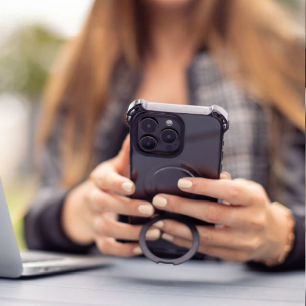 Person holding an iPhone 16 Plus with a Raptic iPhone 16 Shield Impact Case - Dual Impact and a ring grip beside a laptop outdoors, showcasing the case's drop protection features and MagSafe compatibility.