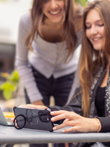 Two women are at a table outside. One, sitting and using a smartphone equipped with the Raptic iPhone 16 Shield Impact Case - Dual Impact for drop protection, is near an open laptop. The other is standing and smiling, bending slightly towards the table.