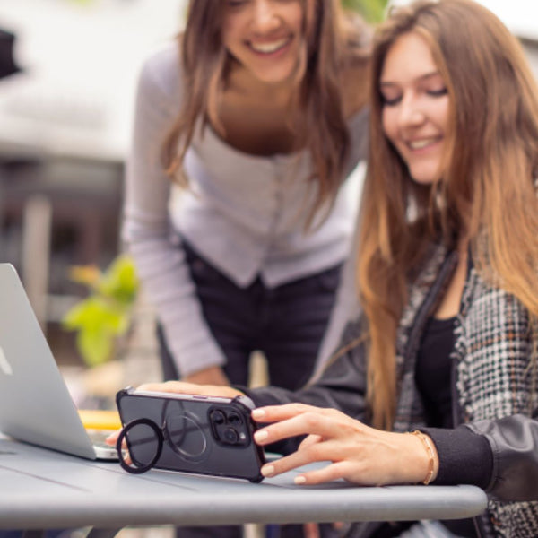 Two women are at a table outside. One, sitting and using a smartphone equipped with the Raptic iPhone 16 Shield Impact Case - Dual Impact for drop protection, is near an open laptop. The other is standing and smiling, bending slightly towards the table.