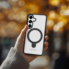 A hand holds a Samsung Galaxy S25 in a white Raptic Air Stand case with a black circular grip, providing military-grade drop protection. Blurred autumn foliage appears in the background.