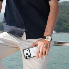 A person in a dark shirt and light pants stands by water, holding a white iPhone in a Raptic iPhone 17 Shield Case with a MagSafe wallet attached; they are wearing a wristwatch.