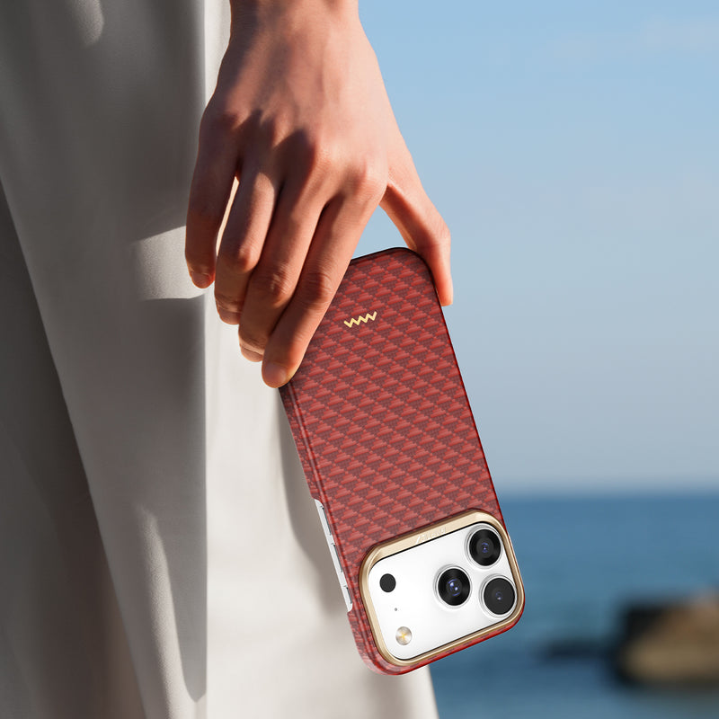 A person stands outdoors near the ocean under a clear sky, holding an iPhone with a red Raptic Skin-Origin Aramid Fibre Case (MagSafe compatible) by Raptic in one hand.