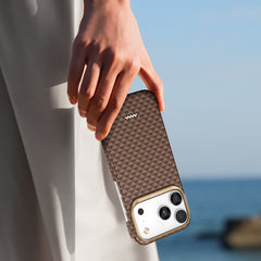 A hand holds an iPhone 17 with the Raptic Skin-Origin ultra-light military-grade aramid fibre case in a brown geometric pattern by the ocean, with part of a light-colored outfit visible.