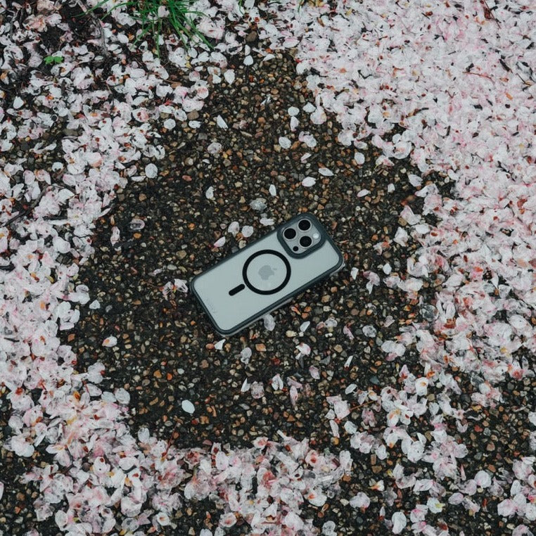 A Raptic iPhone 15 Pro Max Case - Air+ with a dual camera and military spec lies on a ground covered with pink cherry blossom petals.
