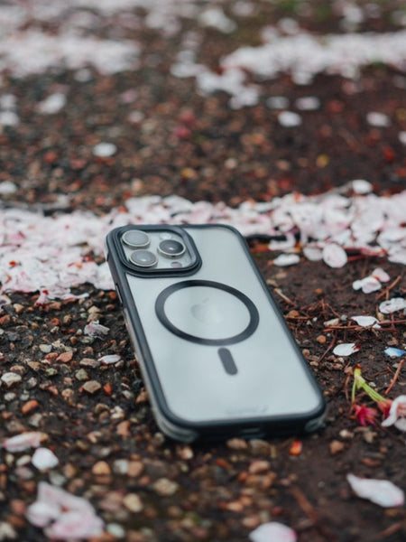 A Raptic iPhone 15 Pro Max case with three camera lenses and military spec drop protection, lying on a gravel pathway scattered with pink cherry blossom petals.
