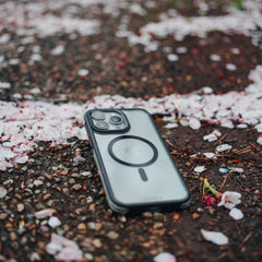 A Raptic iPhone 15 Pro Max case with three camera lenses and military spec drop protection, lying on a gravel pathway scattered with pink cherry blossom petals.