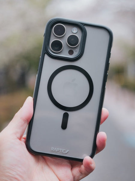 Hand holding a Raptic iPhone 15 Pro Max Case - Air+ with military spec drop protection and camera cutout, with blurred cherry blossoms in the background.
