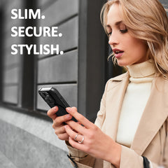 Young woman in a beige trench coat using a Raptic Clear Tough MagSafe Case iPhone 15 - Air 2.0, standing outdoors by a dark wall with the words 