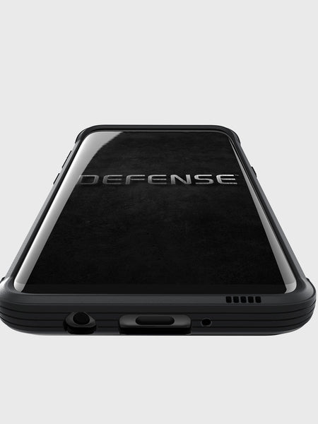 A Samsung Galaxy S8 lies face up on a light grey background, protected by the X-Doria Raptic Lux Black Leather case. The screen displays the word “DEFENSE,” highlighting its military grade drop protection, while the case’s design maintains wireless charging compatibility for everyday convenience.
