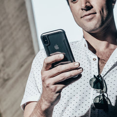 A person confidently holds an iPhone X in a Raptic SHIELD case, renowned for military-grade drop protection, while clutching sunglasses in the other hand and wearing a stylish white patterned shirt.
