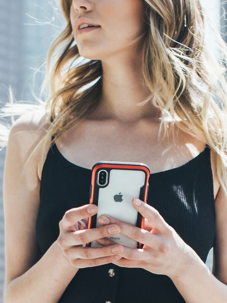 A woman showcasing her iPhone XS in front of a building, protected by a Raptic SHIELD case known for its drop protection.