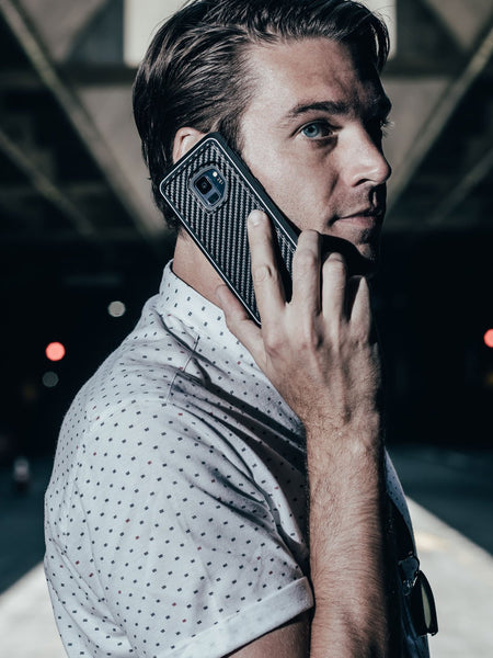 A man in a white patterned shirt holds a smartphone with an X-Doria Samsung Galaxy S9 Case Raptic Lux Black Carbon Fiber to his ear while standing under an overpass at night.
