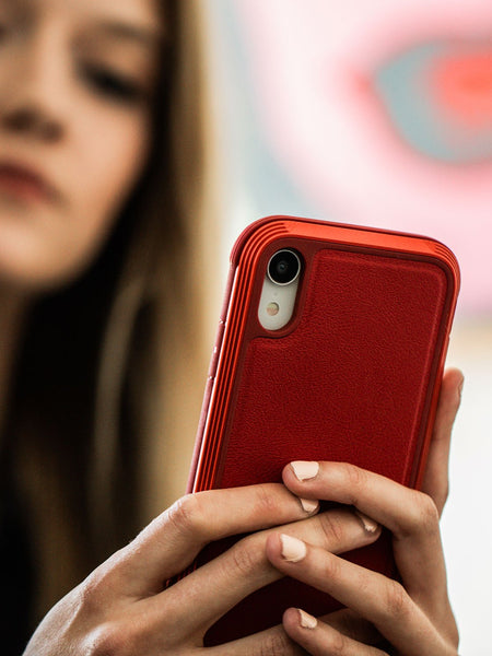 A person is holding a red smartphone, its screen captivating their focus, encased in the impact-resistant polycarbonate LUX case by Raptic for iPhone XR.