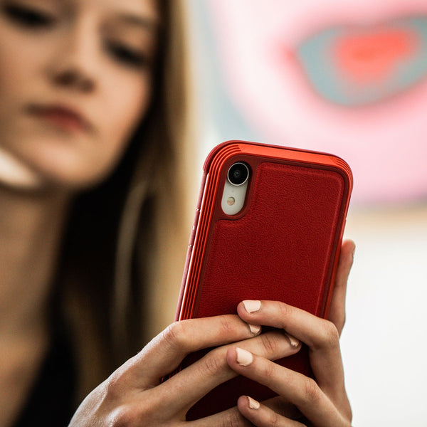 A person is holding a red smartphone, its screen captivating their focus, encased in the impact-resistant polycarbonate LUX case by Raptic for iPhone XR.