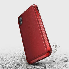 The red iPhone XR Case - LUX by Raptic features a textured back, crafted from impact-resistant polycarbonate. It stands elegantly on a rough grey surface, with small dust particles shimmering around it.